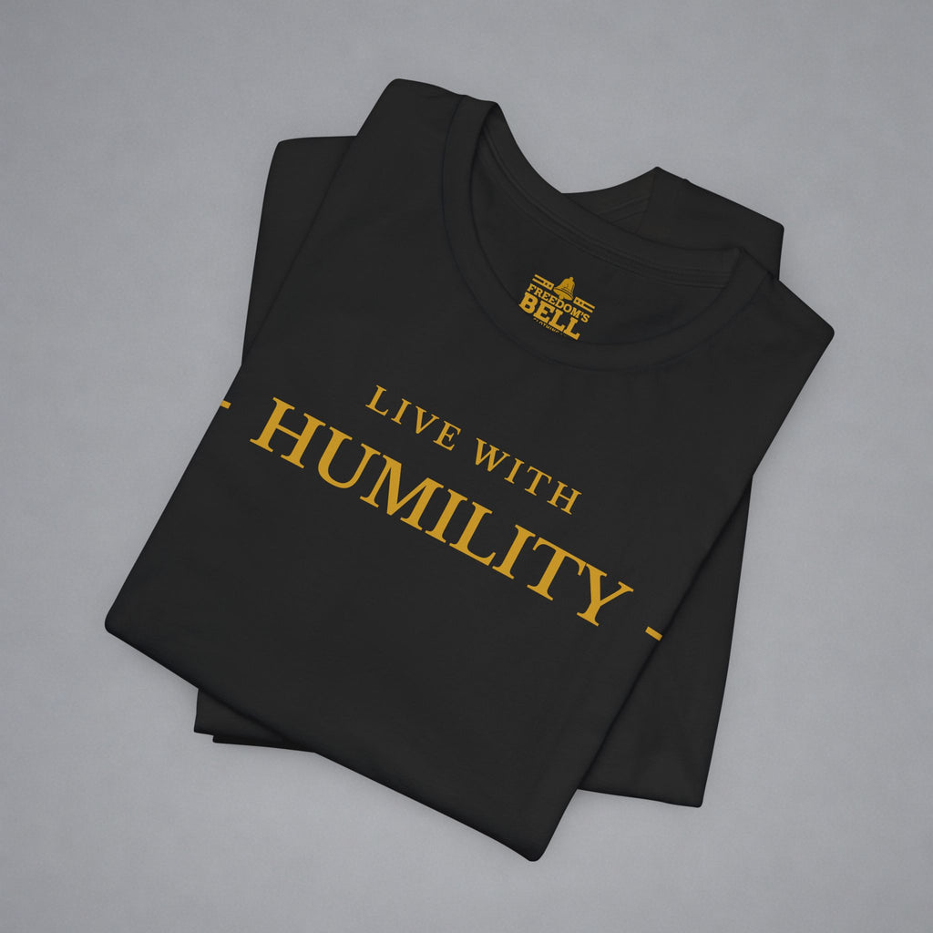 A folded black t-shirt with gold text reading 'LIVE WITH HUMILITY' printed on the front, displayed on a neutral gray background.