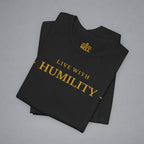 A folded black t-shirt with gold text reading 'LIVE WITH HUMILITY' printed on the front, displayed on a neutral gray background.