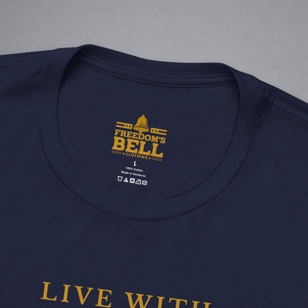 Close-up of a navy blue t-shirt showing the inner neck label with gold text reading 'FREEDOM'S BELL A CLOTHING' and part of the gold 'LIVE WITH' text on the chest, set against a neutral gray background.
