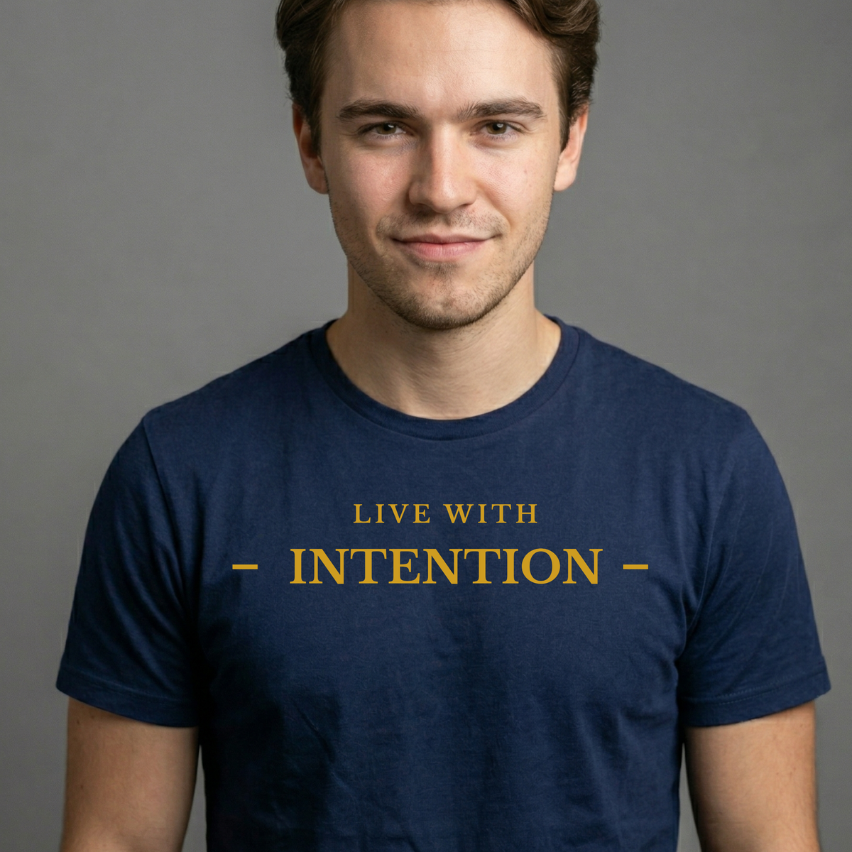 A man wearing a navy blue t-shirt with the text 'LIVE WITH - INTENTION -' printed in gold on the chest, set against a plain gray background.