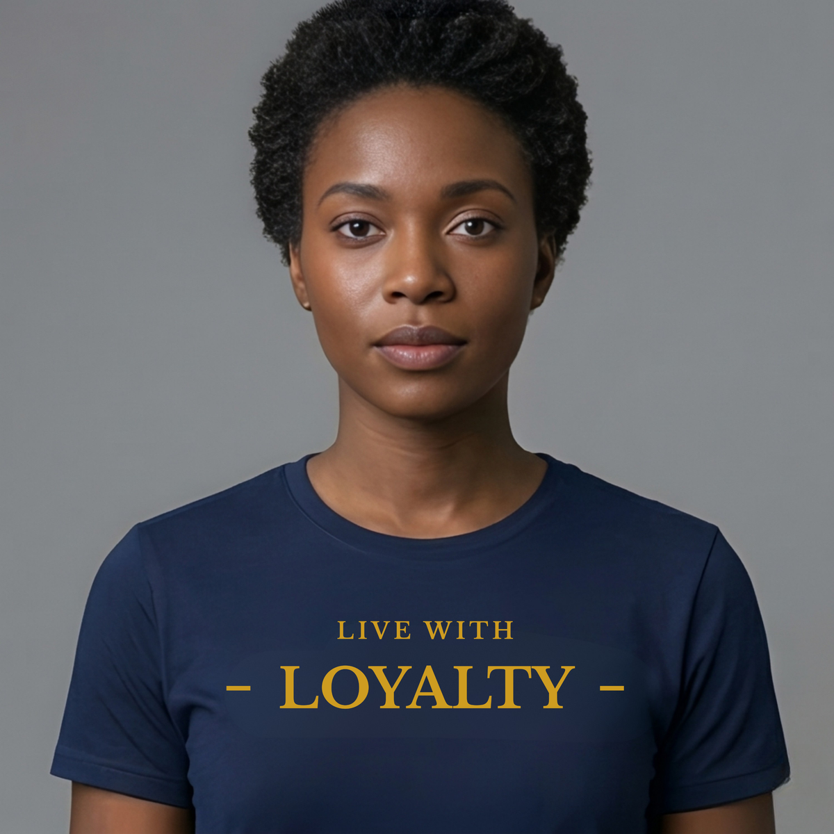 A woman with short curly black hair wearing a navy blue crewneck t-shirt with the text 'LIVE WITH - LOYALTY -' printed in gold on the chest, set against a solid gray background.