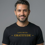A smiling man with short dark hair and a trimmed beard wearing a black crew-neck t-shirt with the words 'LIVE WITH GRATITUDE' printed in gold across the chest, set against a solid gray background.